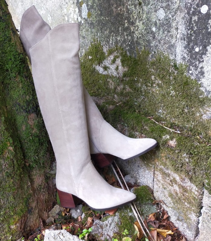 Botas altas de ante beige con tacón medio de madera y puntera afilada, apoyadas sobre un muro de piedra con musgo en un entorno natural otoñal