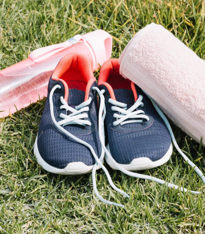 Zapatillas deportivas azul marino con cordones blancos y forro coral, acompañadas de una toalla blanca y una botella rosa sobre césped, imagen de entrenamiento al aire libre