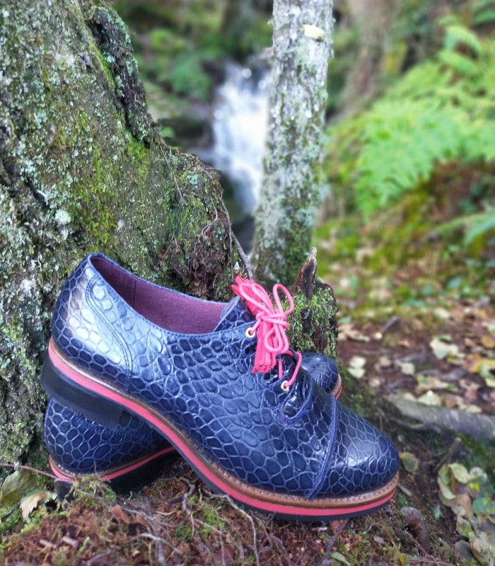 Zapatos de piel con textura cocodrilo en azul marino, cordones fucsia y suela con detalle rojo, fotografiados sobre musgo en un entorno natural de bosque